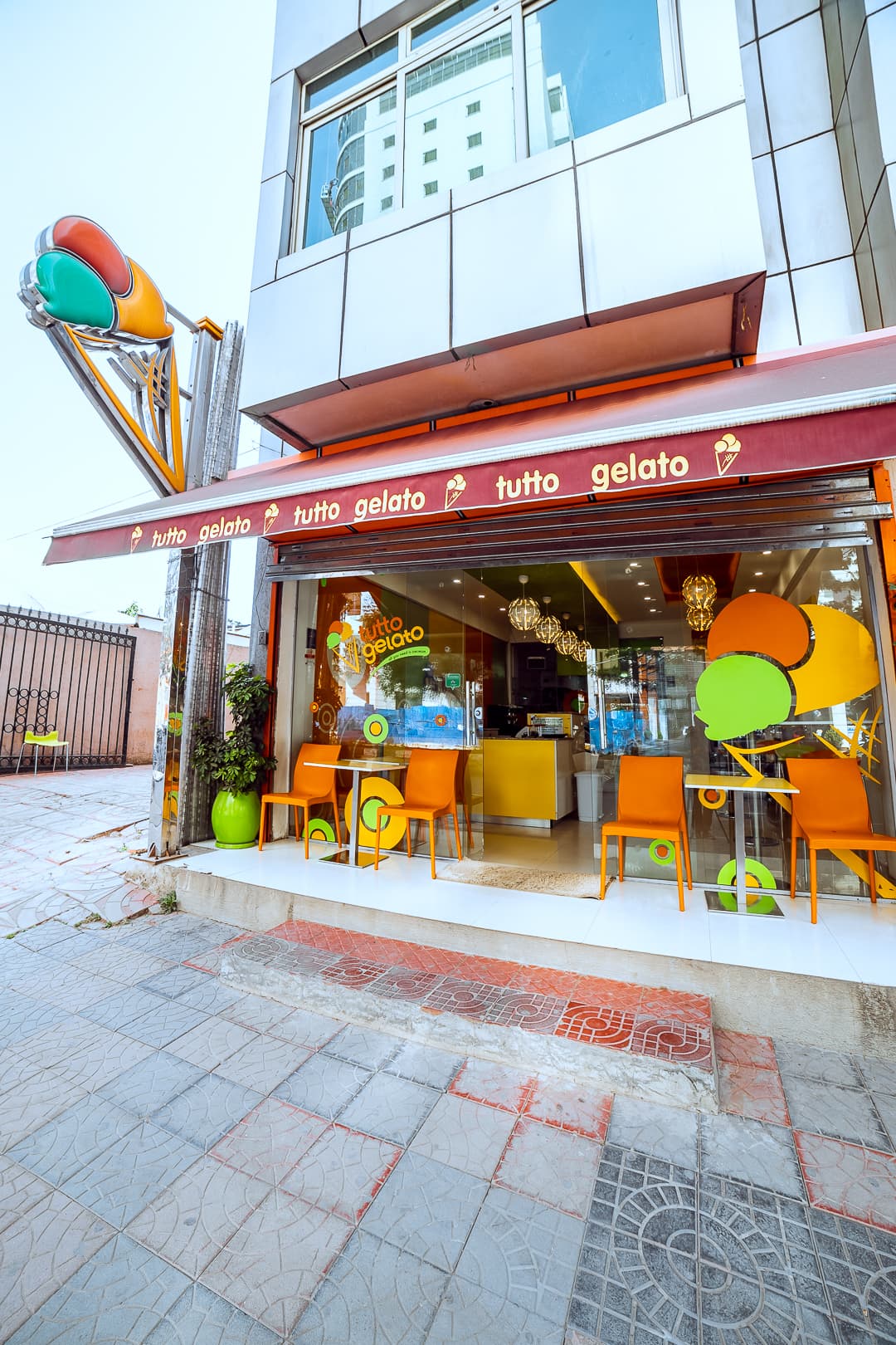 Street perspective of Tutto Gelato store entrance with bright orange outdoor chairs and striped awning